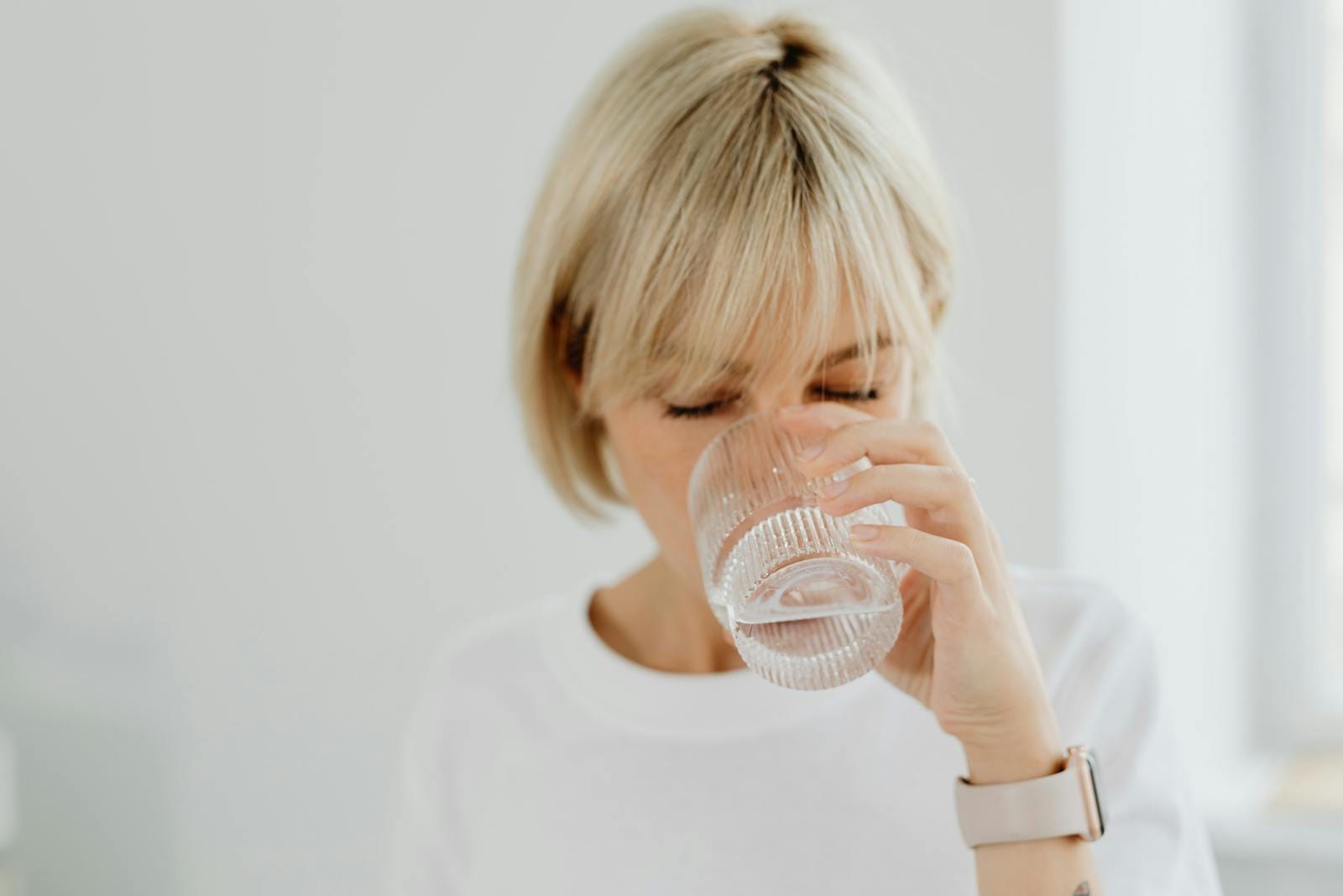 Blonde woman drinking water from glass indoors, showcasing healthy lifestyle and hydration.